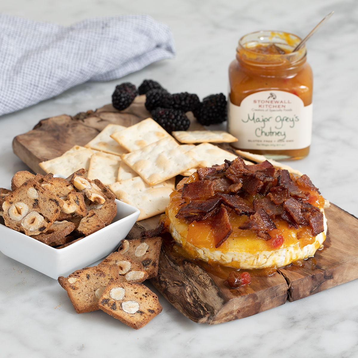 An image of a rustic serving board with a baked brie covered in chutney and bacon pieces flanked by crackers, berries and an open jar of chutney, all on a light gray granite countertop.