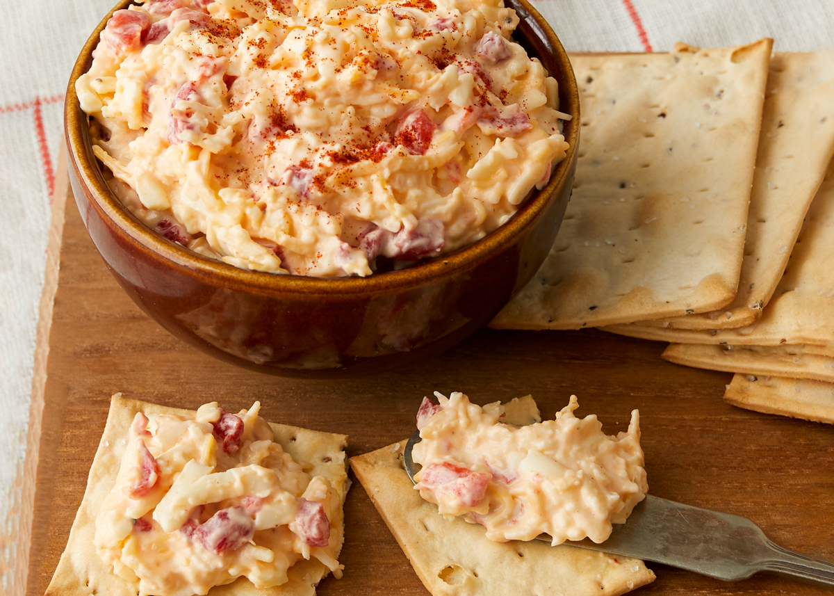 A bowl of Pimento cheese spread in a brown bowl next to some crackers. Some of the crackers have the Pimento spread on them.