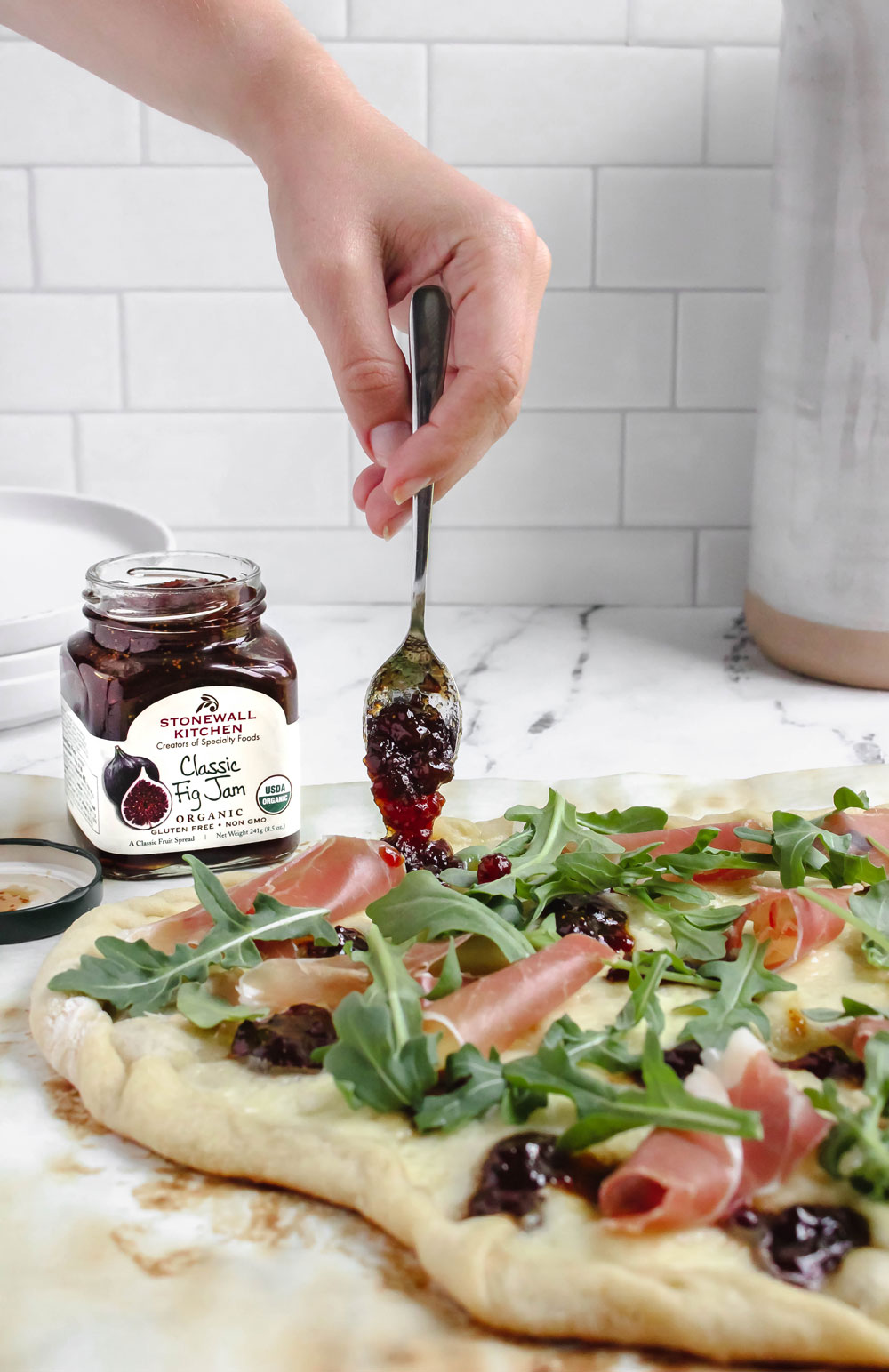 An image of a jar of fig jam next to a prosciutto, brie, and fig jam pizza on a kitchen counter with a white tile backsplash. A hand comes down from the top of the image and scoops some fig jam on the pizza.