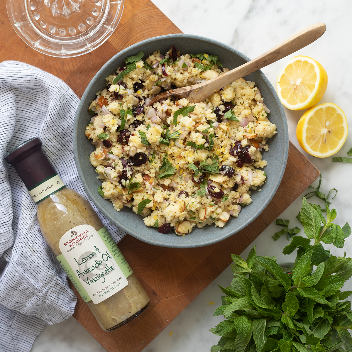 A top-down image of couscous salad in a gray bowl. The bowl is on a wooden serving tray and it is surrounded by a number of things like a bottle of vinaigrette, mint sprigs, cut lemons and a light blue napkin.