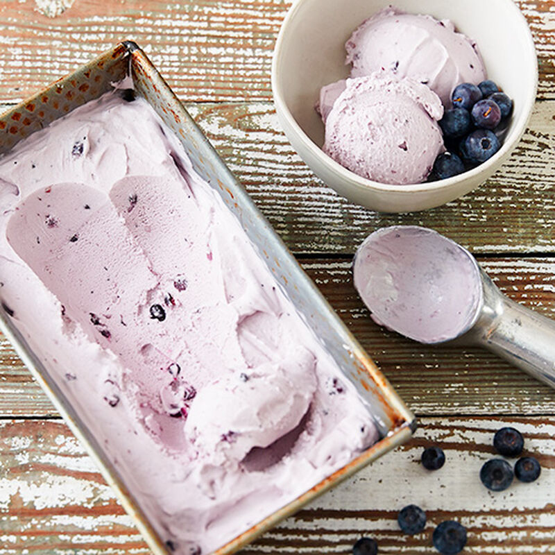 image of a rectangular tin filled with light purple blueberry ice cream with an ice cream scoop and a bowl of freshly scooped ice cream next to it