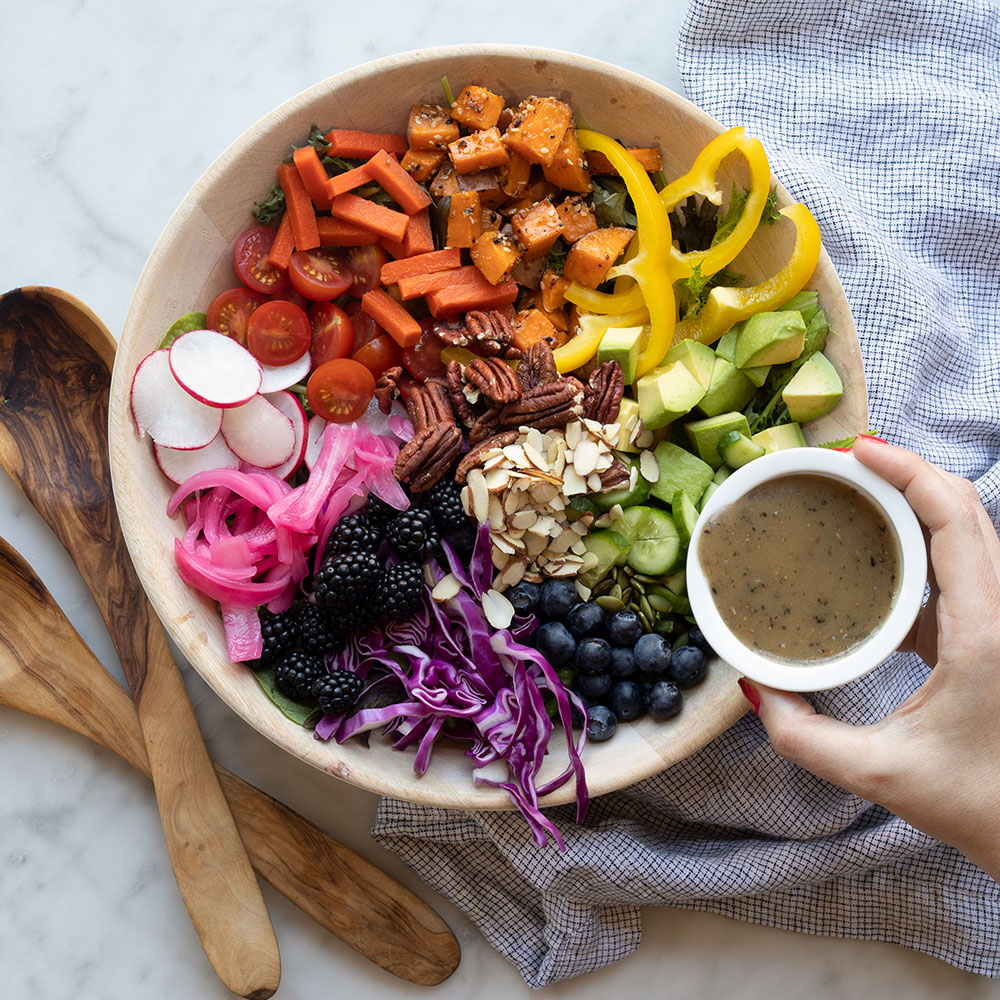 image of a bowl with salad in it, arranged to look like a rainbow and there is a hand with a small jar of dressing that is ready to pour over the dish