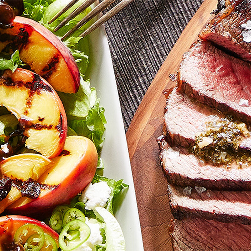 image of a plate of grilled peaches next to a wooden cutting board with chimichurri steak thinly cut and arrayed on it