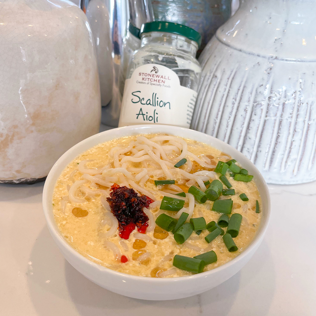 An image of a white bowl of ramen garnished with green onions and chilis. An empty jar of Scallion Aioli is behind the bowl and is in between two large ceramic jars.