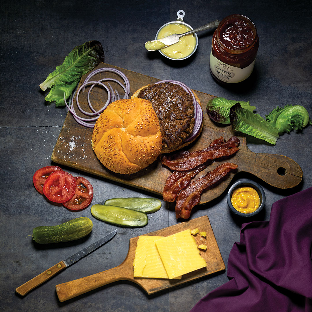 An image of a rustic serving board with a baked brie covered in chutney and bacon pieces flanked by crackers, berries and an open jar of chutney, all on a light gray granite countertop.