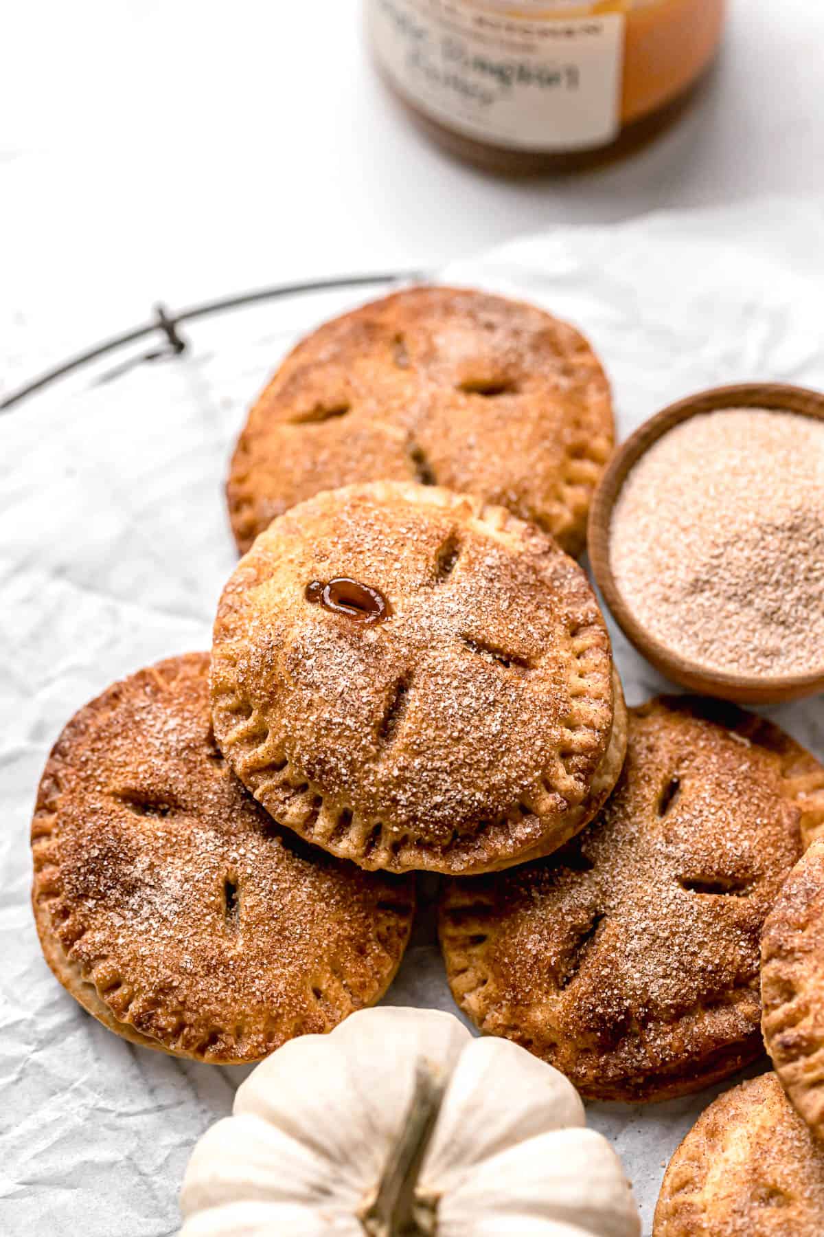 a pile of baked hand pies with filling oozing out on a counter next to a bowl of cinnamon sugar and a white pumpkin
