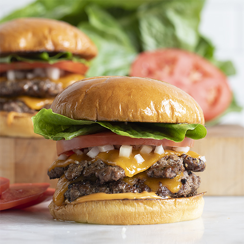 two smashburgers on a counter with tomatoes and lettuce in the background