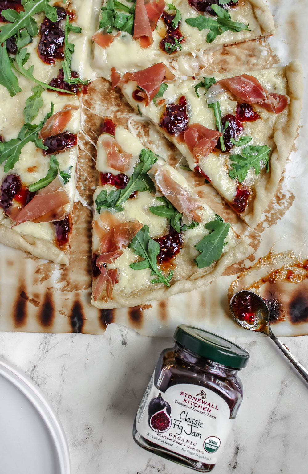 A top-down image of a jar of fig jam next to a prosciutto, brie, and fig jam pizza on a kitchen counter.