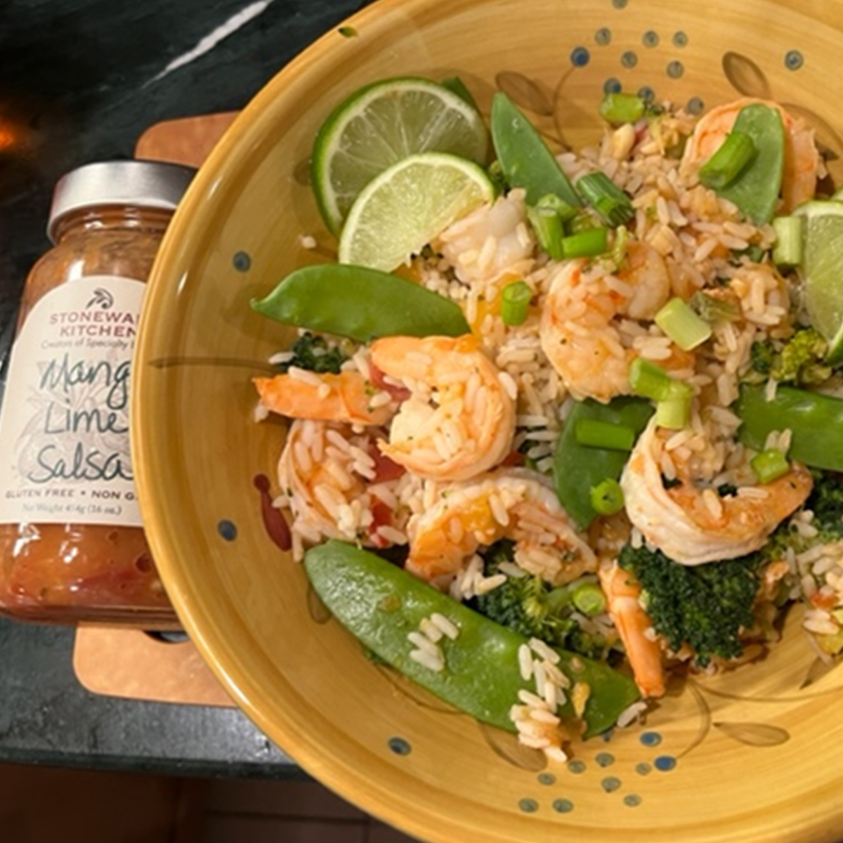 On a dark countertop, a large yellow bowl is filled with a garlicky shrimp dish, with rice, limes, and peas mixed throughout. To the left of the bowl is a jar of Mango Lime Salsa.