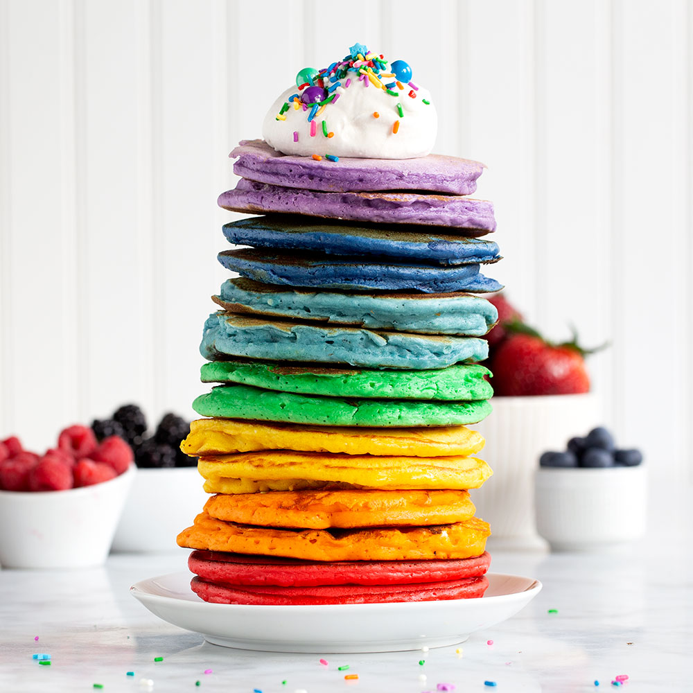 image of a stack of rainbow pancakes on a counter with whipped cream and sprinkles on top