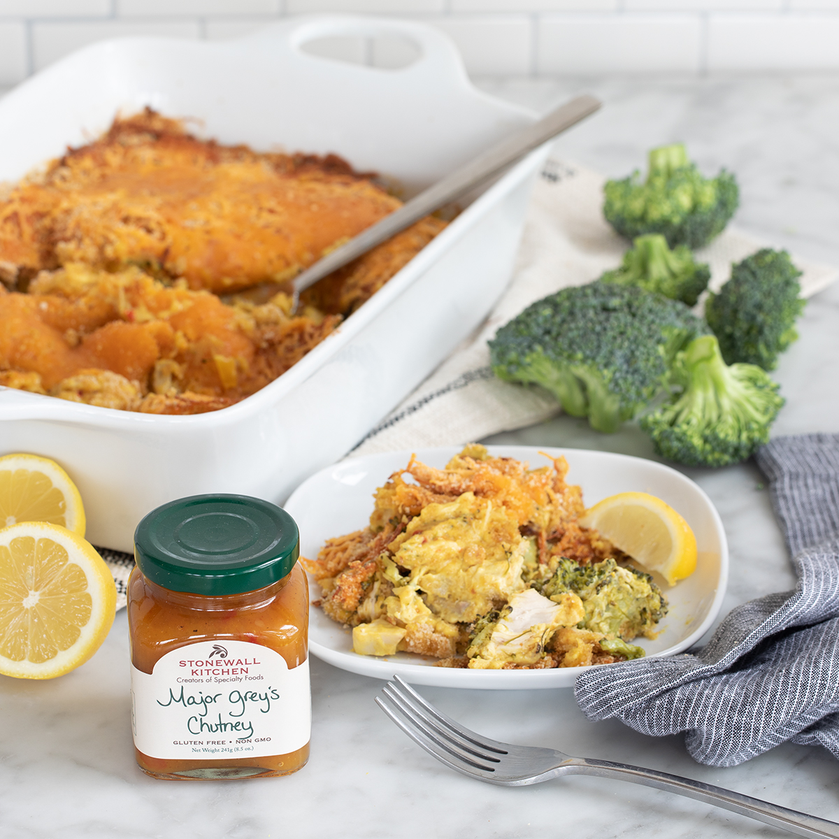 An image of a small plate of Chicken Divan with a lemon slice garnish. A white casserole dish is in the background with more of the chicken in it. A jar of chutney is in the foreground and behind the plate is some florets of broccoli.