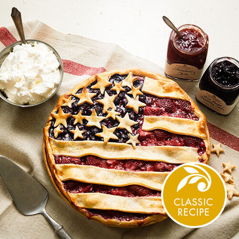 image of the stars and stripes pie near a bowl of whipped cream and displayed on a rustic, flag-like napkin with two open jars of Stonewall Kitchen jams